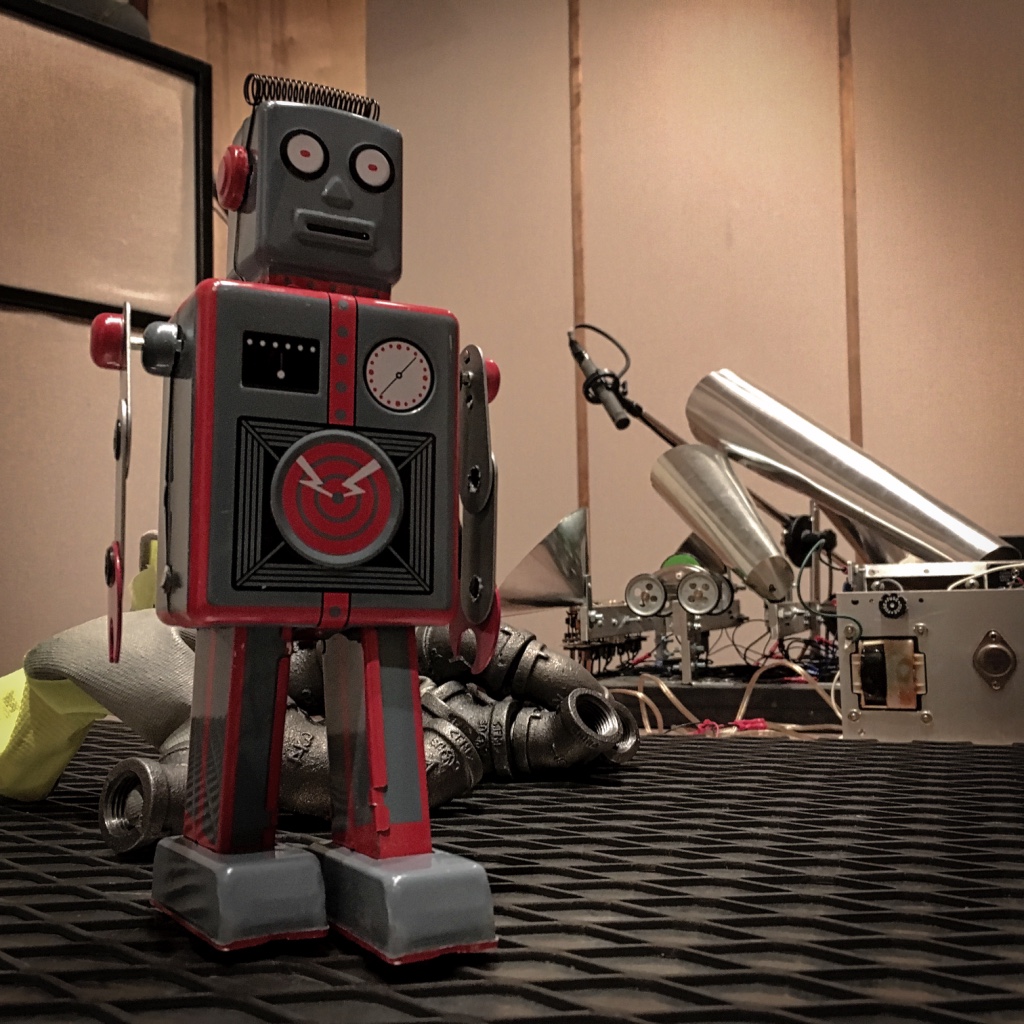 Toy robot standing on a metal work surface with audio equipment behind it.