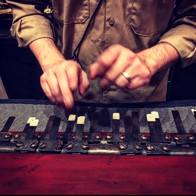 Close-up of hands playing a custom mechanical soundmaking instrument with metal keys and dampers.