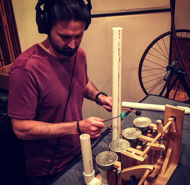 Person wearing headphones striking a custom-built soundmaking device with mallets in a studio.